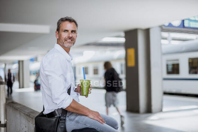Pensativo hombre de negocios sosteniendo la bebida mirando hacia otro lado mientras está sentado en la plataforma de la estación de tren - foto de stock