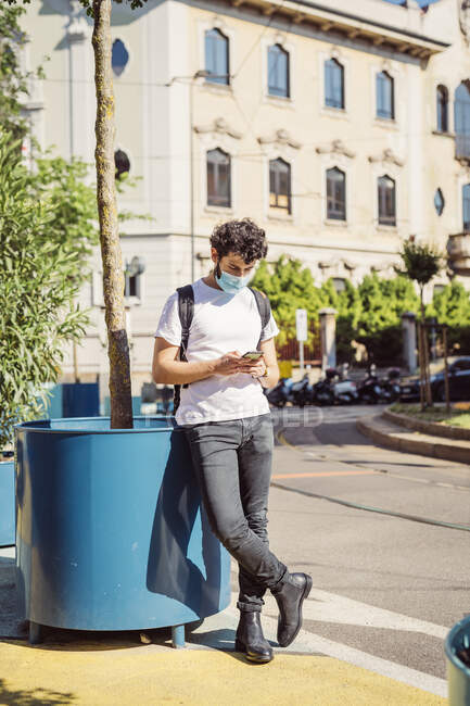 Hombre usando máscara usando teléfono móvil mientras está parado en la acera en la ciudad - foto de stock