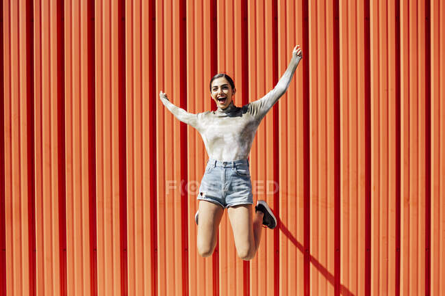Full length of carefree young woman jumping with arms outstretched against red corrugated wall on sunny day — Stock Photo