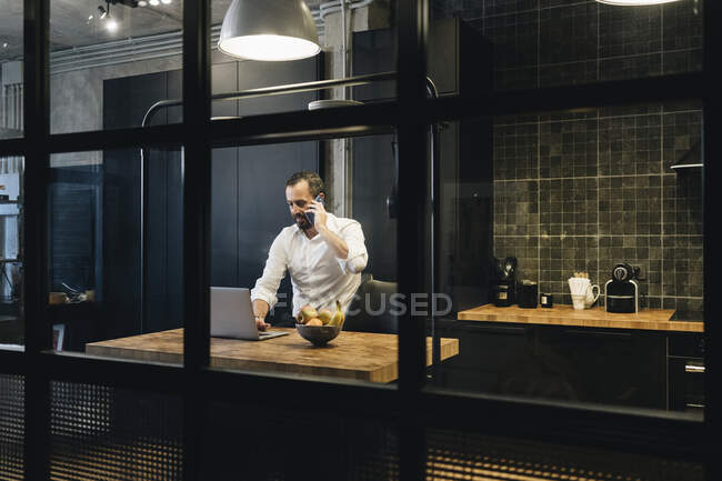 Homme d'affaires mature travaillant dans la cuisine, utilisant un ordinateur portable, parlant au téléphone — Photo de stock