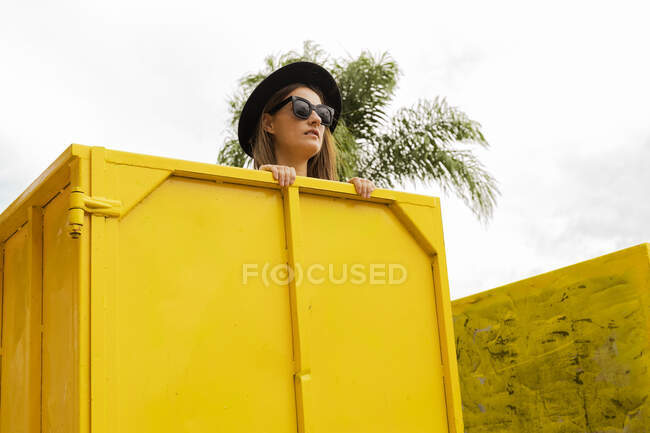 Жінка з сонцезахисними окулярами дивиться через край жовтого контейнера. — стокове фото