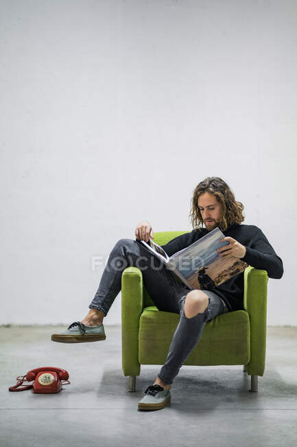 Junger Mann liest Buch, während er zu Hause auf Sessel gegen Wand sitzt — Stockfoto