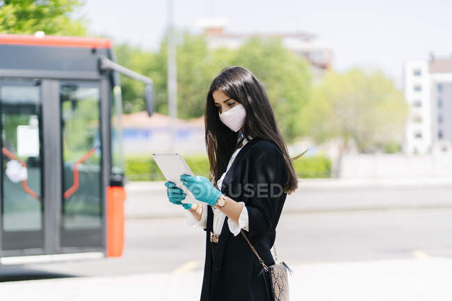 Junge Frau mit Schutzmaske und Handschuhen mit digitalem Tablet beim Warten an Bushaltestelle, Spanien — Stockfoto