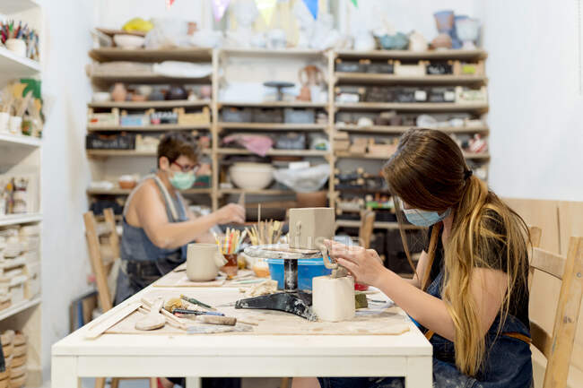 Mujeres con máscaras haciendo cerámica en el banco en el taller - foto de stock