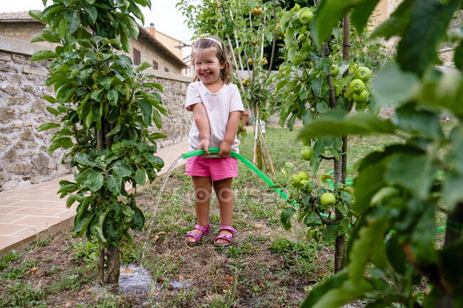 Смеющаяся маленькая девочка поливает дерево в саду — стоковое фото