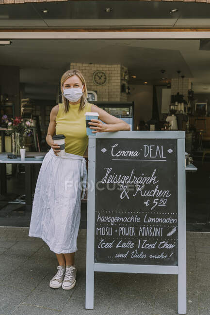 Femme portant un masque facial et tenant du café à emporter tout en s'appuyant sur un panneau avec des offres spéciales Corona — Photo de stock
