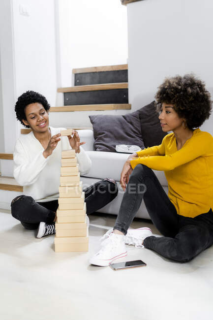 Mujer joven sonriente apilando cajas en el suelo en casa mientras su amiga la observa - foto de stock