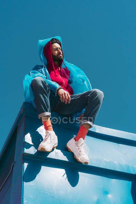 Молодий чоловік сидить на краю блакитного контейнера в дощовому пальто. — стокове фото