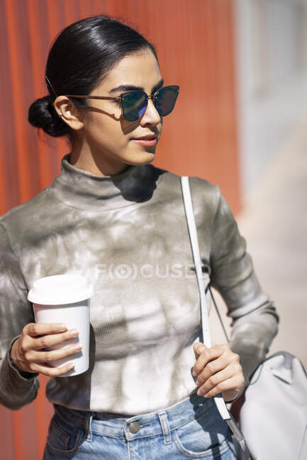 Mujer joven de moda sosteniendo la taza de café mientras mira hacia otro lado en el día soleado - foto de stock