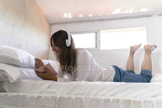 Усміхнена жінка лежить на ліжку і слухає музику з навушниками під час нотаток. — стокове фото