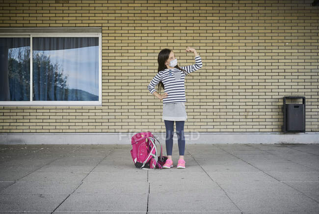 Девушка в школьной сумке в маске стоит перед зданием, напрягая мышцы — стоковое фото