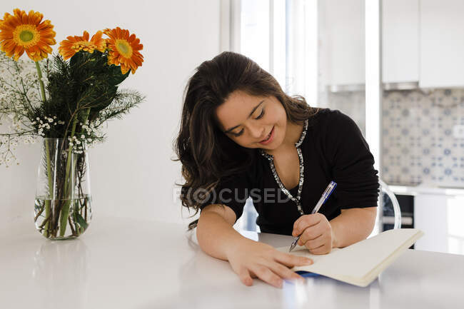 Giovane donna felice con sindrome di Down scrivere in libro da vaso di fiori sul tavolo a casa — Foto stock