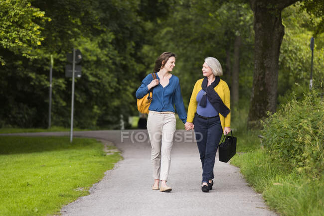 Sorridente donna anziana e figlia adulta che camminano mano nella mano in un parco guardandosi l'un l'altro — Foto stock