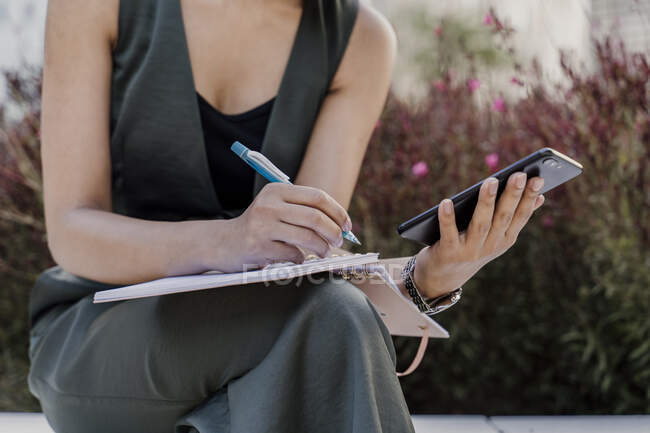Close-up of businesswoman holding smart phone writing in note pad while sitting outdoors — Stock Photo
