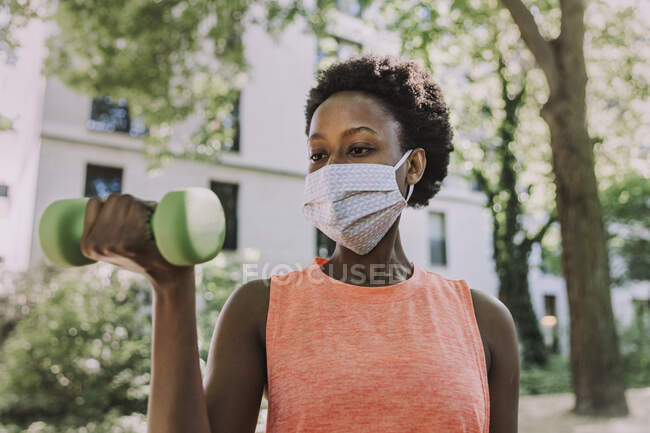 Retrato de mulher usando máscara protetora exercitando com halteres ao ar livre — Fotografia de Stock