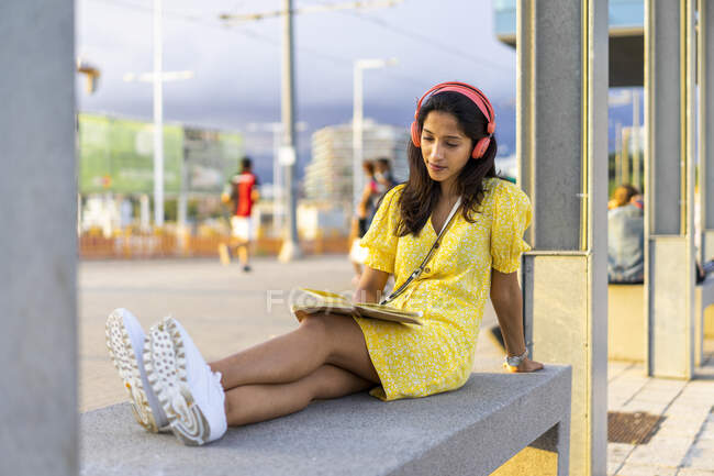 Молодая женщина слушает музыку во время чтения книги о бетонной скамейке в городе — стоковое фото