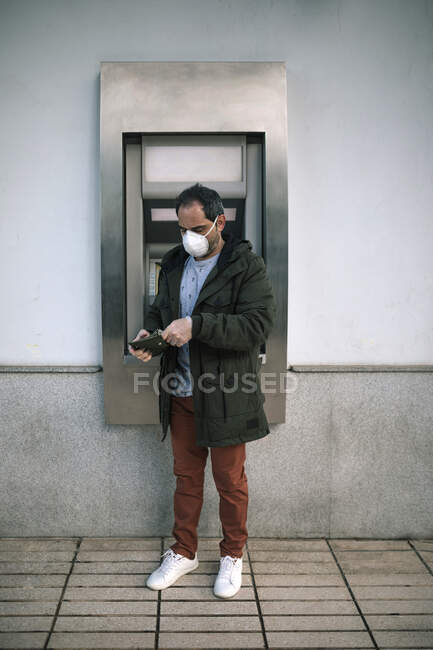 Hombre con máscara protectora de pie frente al cajero automático poniendo dinero en su bolso - foto de stock