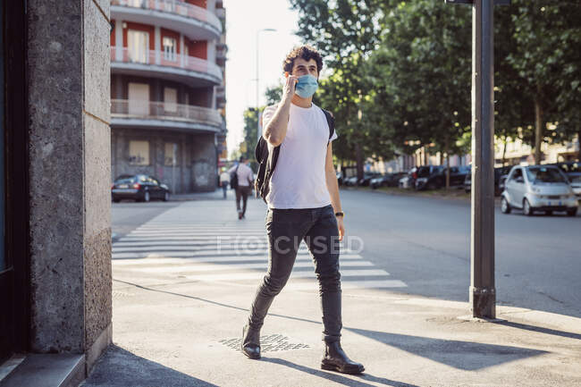 Mann mit Gesichtsmaske spricht über Smartphone, während er in der Stadt auf der Straße läuft — Stockfoto