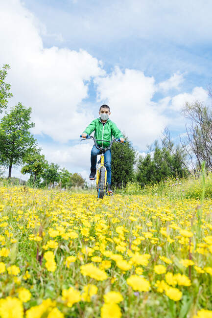Хлопчик, одягнений в маску обличчя, їздить по жовтих квітах на полі проти неба. — стокове фото