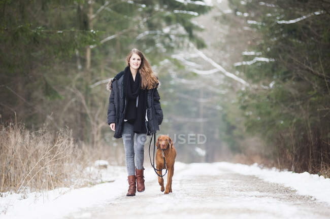 Beautiful young woman walking with dog on road amidst trees in forest during winter — Stock Photo