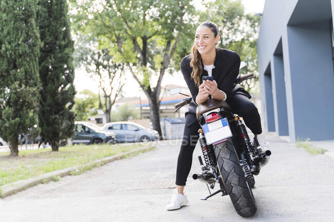 Mujer sonriente sentada en moto y usando smartphone - foto de stock