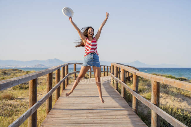 Emocionado adolescente saltando en el paseo marítimo en la playa contra el cielo despejado - foto de stock