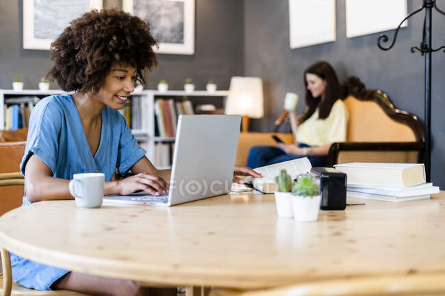 Donna felice utilizzando il computer portatile al caffè con un amico in background — Foto stock