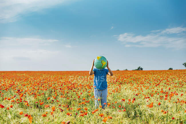 Pré-adolescent garçon tenant globe tandis que debout dans le champ de pavot contre le ciel bleu — Photo de stock