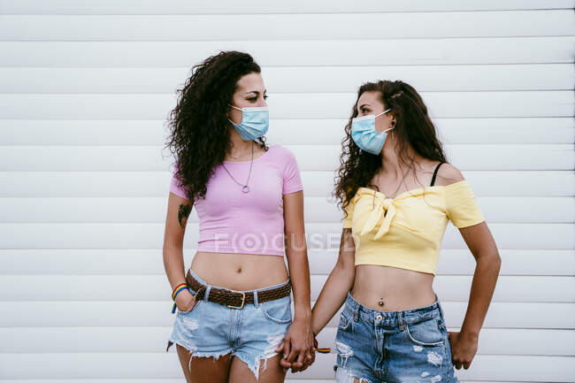 Lésbicas casal vestindo máscaras de mãos dadas enquanto de pé contra a parede na cidade — Fotografia de Stock