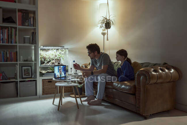 Niña enferma sentada por el padre discutiendo la telemedicina con el médico sobre la videollamada a través del ordenador portátil en la sala de estar en casa - foto de stock