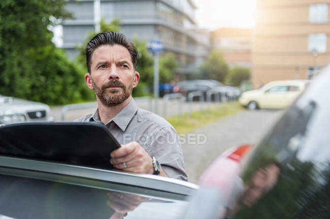 Pensativo hombre de negocios mirando hacia otro lado mientras sostiene archivo en el techo del coche en el estacionamiento en la ciudad - foto de stock
