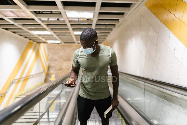Jeune homme portant un masque à l'aide d'un téléphone intelligent debout sur l'escalator à la station de métro — Photo de stock