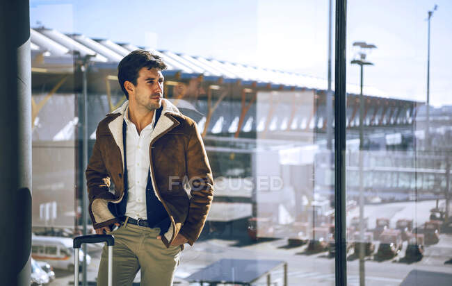 Thoughtful businessman standing with hands in pockets by window at airport departure area — Stock Photo