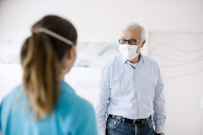 Uomo anziano che incontra il suo fisioterapista femminile alla reception, indossando maschere per il viso — Foto stock