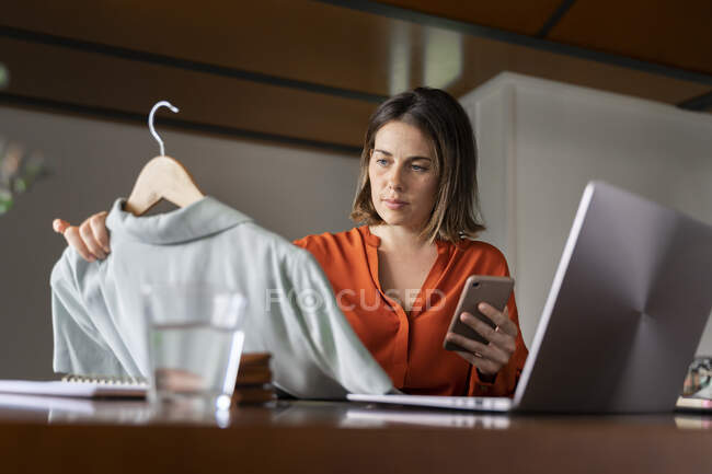 Businesswoman with smart phone looking at clothes while sitting at home — Stock Photo