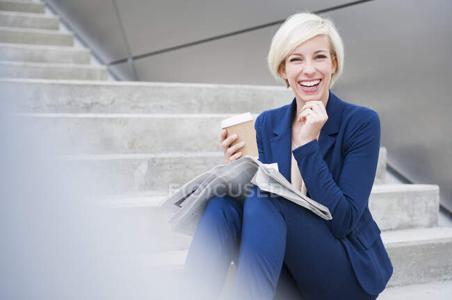 Portrait of blond laughing businesswoman with coffee to go and newspaper sitting on stairs — Stock Photo