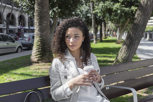 Задумчивая стильная деловая женщина держит чашку кофе, сидя на скамейке в парке — стоковое фото