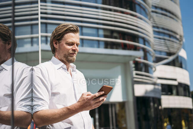 Businessman in the city leaning against glass front checking smartphone — Stock Photo