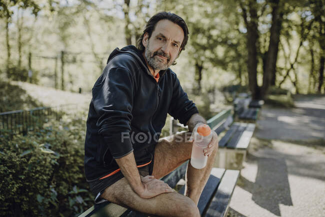 Homme mature confiant tenant une bouteille d'eau tout en étant assis sur le banc dans le parc — Photo de stock