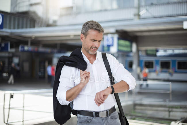 Homme d'affaires tenant blazer vérifier le temps tout en se tenant contre la gare — Photo de stock
