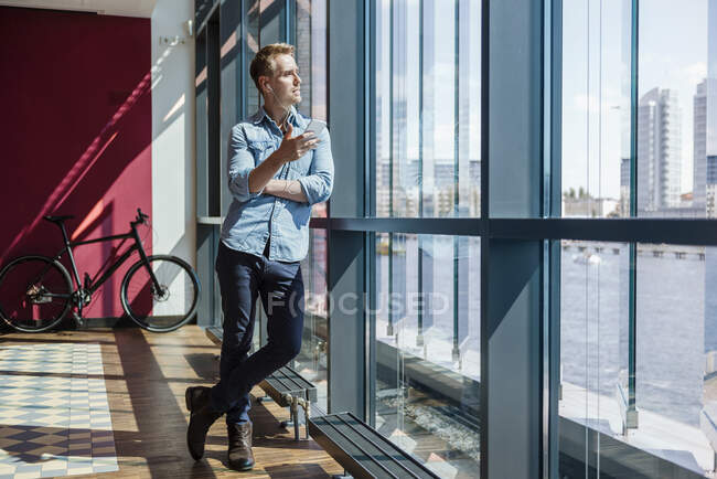 Hombre de negocios casual de pie en la oficina moderna, utilizando teléfonos inteligentes y auriculares - foto de stock