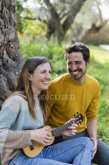Cheerful man looking at girlfriend playing guitar while sitting in countryside — Stock Photo