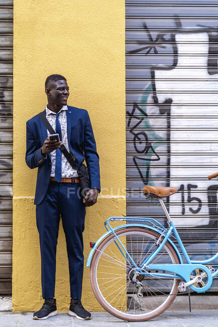 Joven hombre de negocios sonriente con smartphone y bicicleta en la ciudad - foto de stock