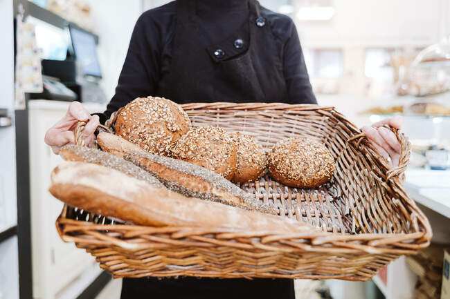 Женщина пекарь держит различные хлеба в плетеной корзине в пекарне — стоковое фото