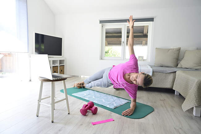 Reife Frau mit Laptop praktiziert Yoga auf Turnmatte im Wohnzimmer — Stockfoto