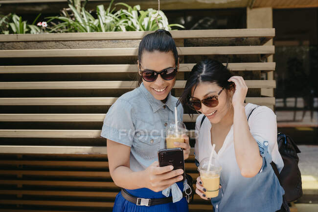 Amigas usando gafas de sol mirando fotografía por teléfono inteligente en la ciudad - foto de stock
