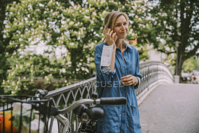 Mujer en un puente con máscara facial y bicicleta hablando por teléfono - foto de stock