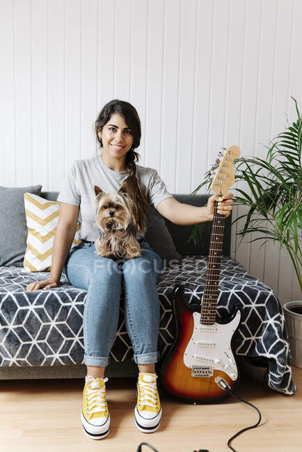 Jovem feliz segurando guitarra elétrica enquanto se senta com Yorkshire Terrier no sofá em casa — Fotografia de Stock