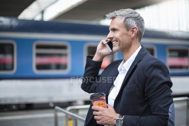 Бізнесмен тримає каву, розмовляючи по смартфону, стоячи на залізничній станції — стокове фото