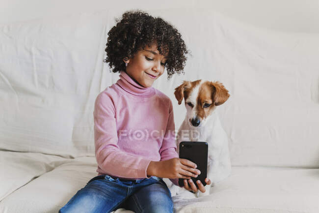 Porträt eines lächelnden kleinen Mädchens, das mit seinem Hund auf der Couch sitzt und ein Selfie mit dem Smartphone macht — Stockfoto